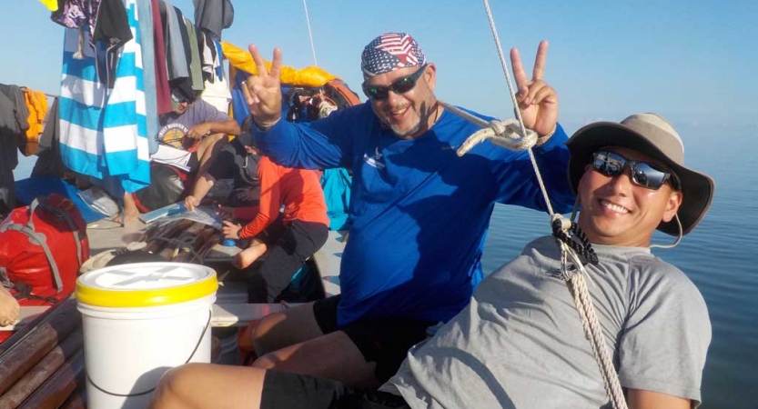 Two people smile while sitting on a boat. One of them gives the camera peace signs. 